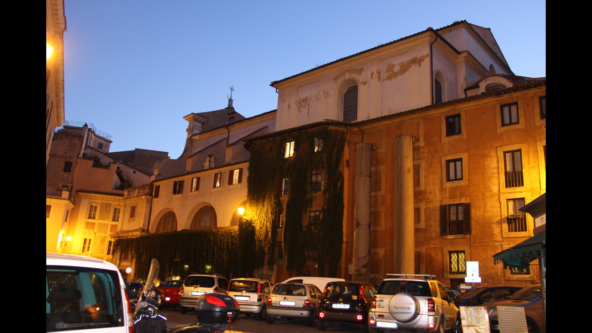 Un bâtiment imposant éclairé en soirée, avec des ouvertures régulières et une façade partiellement recouverte de végétation grimpante.