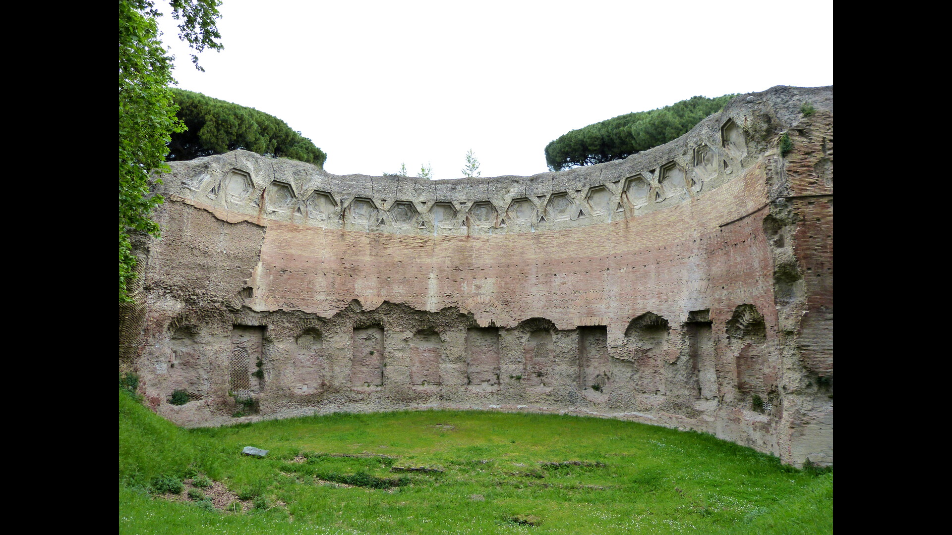 Un mur circulaire ancien en pierre et brique, partiellement érodé, avec des ouvertures rectangulaires et des niches vides alignées sur sa partie supérieure.